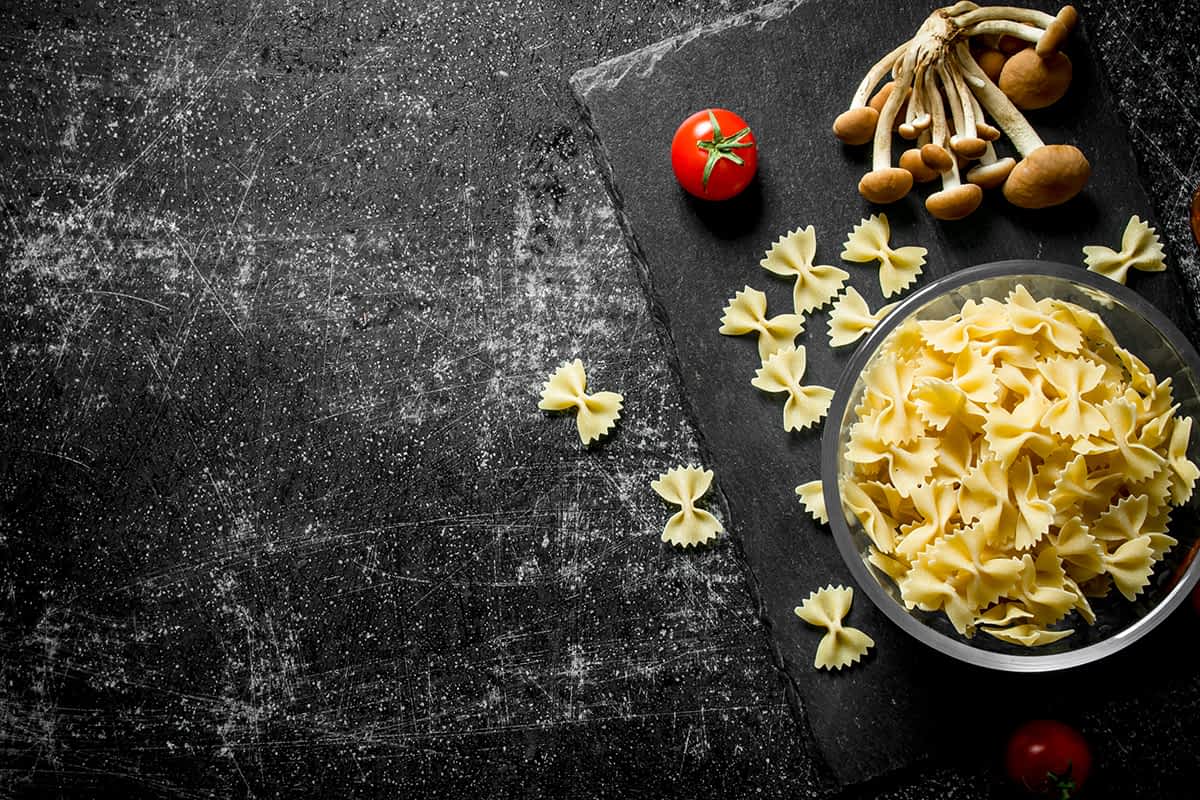 Raw Farfalle pasta in a bowl with mushrooms and tomatoes. On black rustic background