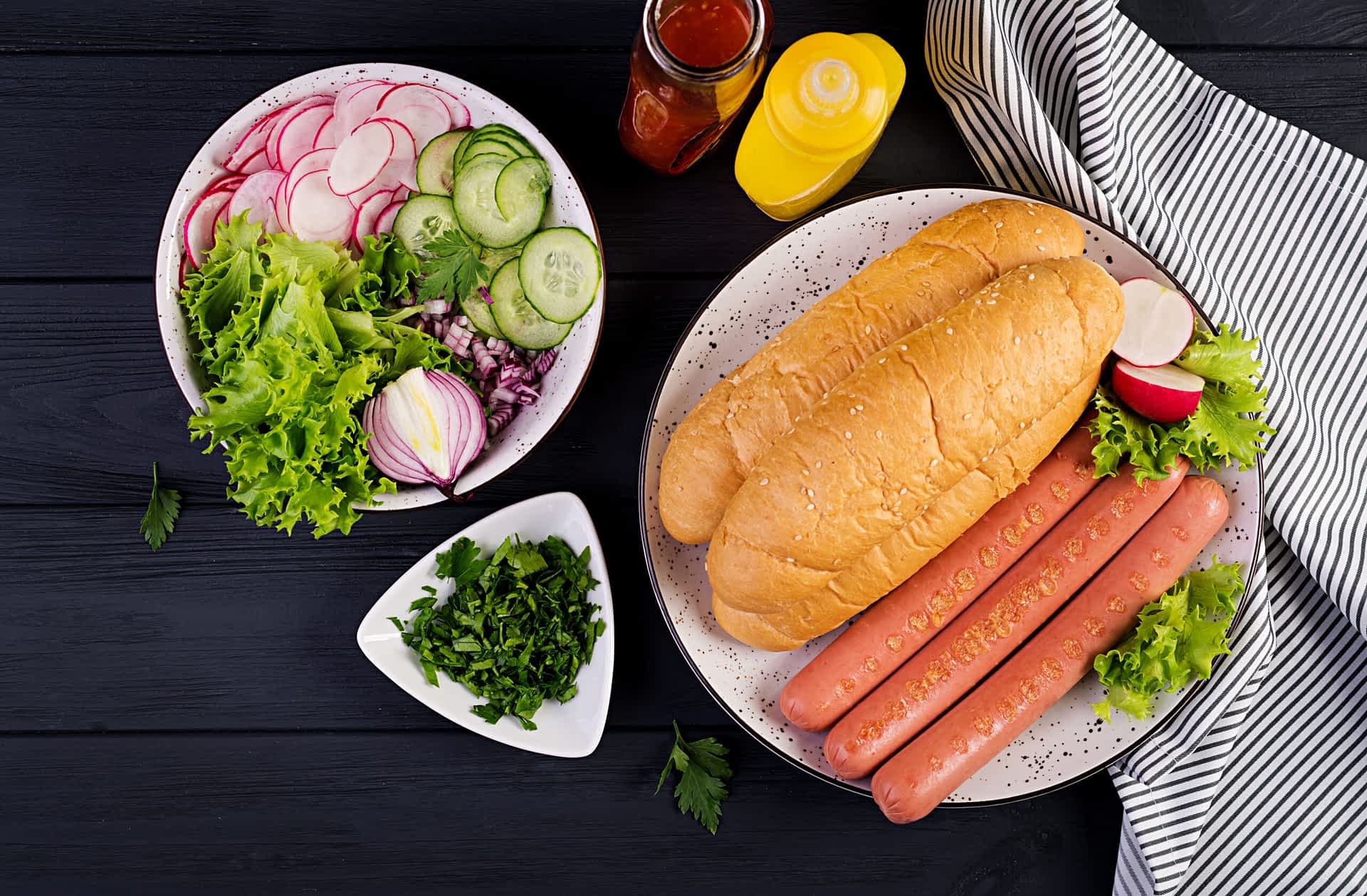 Ingredients for hot dog with  sausage, cucumber, radish and lettuce on dark wooden background. Summer hotdog. Top view