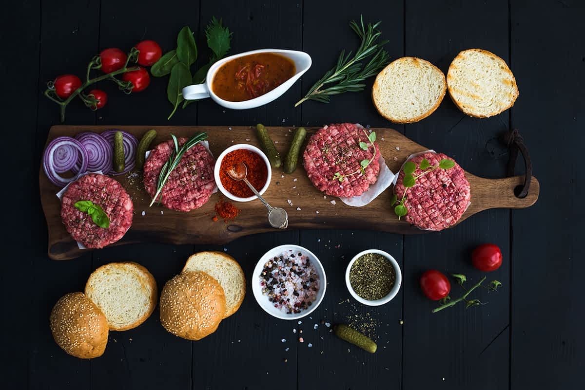 Ingredients for cooking burgers. Raw ground beef meat cutlets on wooden chopping board, red onion, cherry tomatoes, greens, pickles, tomato sauce, cheese, herbs and spices over black background, top view, horizontal
